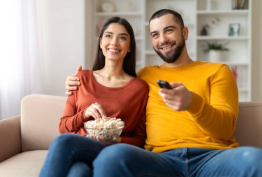 Eve. Rahatla. Mutlu Genç Evli Çift Oturma Odasında TV izliyor, Milenyum Eşleri birlikte patlamış mısır yiyor, rahat koltukta dinleniyor, Uzaktan kumandalı adam, yakın çekim...