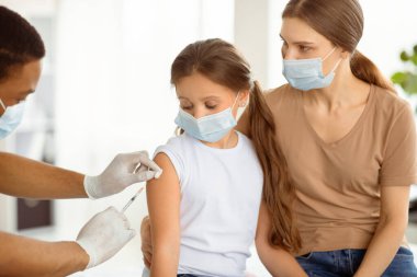 Maskeli ebeveyn ve çocuk koronavirüse karşı aşı oluyor, doktor klinikte kovid 19 aşısı yapıyor. Anne ve kızı aşı kampanyasına katılıyor.