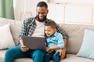 Afrikalı baba kucağında dizüstü bilgisayarla oturmuş bilgisayarda çizgi film seyrediyor birlikte vakit geçiriyor ve evde kaynaşıyor. 