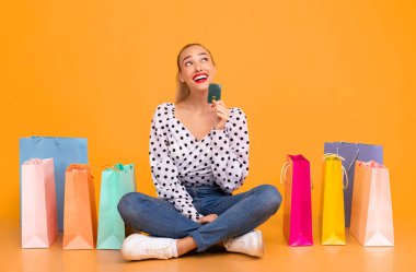 Ne alacağımı. Kredi kartı tutan trend bir kadının portresi ve fotokopi odasına bakmak, satın almayı düşünmek, yeni kıyafetler hayal etmek, sarı stüdyo arka planında yerde oturmak.