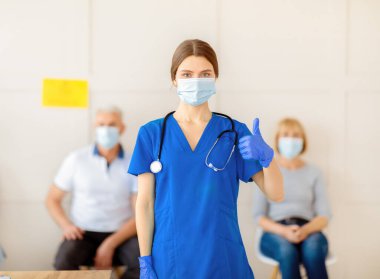 Yüz maskeli, eldivenli ve üniformalı genç bir bayan doktor. Klinikte kovid 19 'a karşı bağışıklık kazanmayı öneriyor. Koronavirüs aşısı için bekleyen kıdemli hastalar