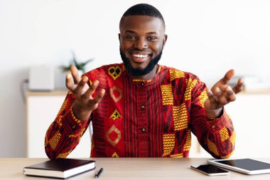 Gülümseyen Afrikalı bir adamın portresi ofis masasında oturuyor, kameraya konuşuyor, geleneksel kıyafet giymiş neşeli siyahi danışman iş deneyimini ve iş fikirlerini paylaşıyor, bedava alan.