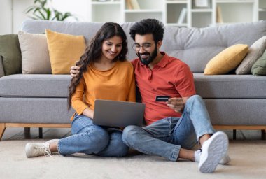 E-ticaret kavramı. Bilgisayarlı ve kredi kartlı mutlu Hintli çift evlerinden online alışveriş yapıyorlar, gülümseyen Doğu eşleri oturma odasında bilgisayar başında oturuyorlar, internet alışverişi yapıyorlar.