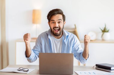 Ofiste dizüstü bilgisayarla başarıyı kutlayan neşeli Doğulu adamın portresi, heyecanlı Arap adam iyi habere duygusal tepki veriyor, yumruklarını kaldırıyor ve Joy 'la ünleniyor, terfi alıyor, boşluğu kopyalıyor.