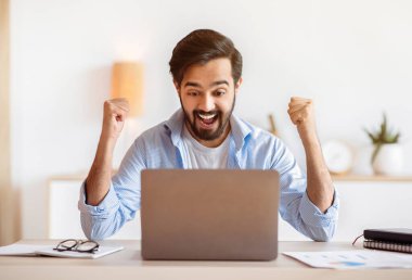 Heyecanlı Arap adam başarıyı ev bilgisayarıyla kutluyor, iyi haberlere duygusal tepki veriyor, masa başında oturuyor, bilgisayar ekranına bakıyor, yumruklarını kaldırıyor ve heyecanla bağırıyor.