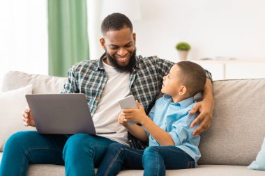 Afrika kökenli Amerikalı baba ve oğul akıllı telefon ve dizüstü bilgisayar kullanarak evdeki koltukta oturuyorlar. Mobil Uygulama. Aile Gadget 'ları, bilgisayar ve telefon kullanımı, evdeki interneti tarama. Seçici Odaklanma