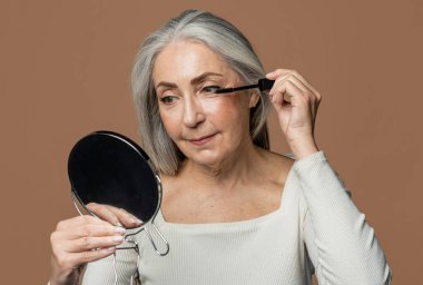 Neşeli son sınıf beyaz kadın, gri saçlı, kirpiklerine rimel sürüyor, aynaya bakıyor, kahverengi arka planda, stüdyoda izole edilmiş. Yaşam tarzı, güzellik, doğal makyaj, günlük kozmetik.