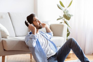 Kablosuz kulaklıklı genç Arap adam evde müzik dinlemekten zevk alıyor, Milenyum Doğulu adam oturma odasında gözleri kapalı oturuyor, favori melodileri ile dinleniyor,