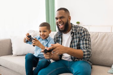 Heyecanlı Afrikalı baba ve oğlu evdeki koltukta oturup eğleniyorlar. Genç Siyah Baba ve Oğlu Hafta Sonu Konsepti 'nde birlikte vakit geçirerek video oyunları oynuyorlar.