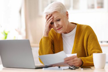 Son sınıf öğrencisi kadın faturaları okurken başını dik tutuyor, karantina sırasında maddi sorunlar yaşıyor. Mutfakta dizüstü bilgisayarın önünde oturan üzgün yaşlı kadın ve elinde belgeler, boşluğu kopyala