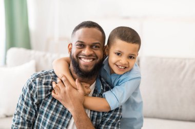 Neşeli Afrikalı baba ve oğulun evdeki koltukta oturmuş kameraya gülümseyen pozu portresi. Mutlu Babalık ve Çocukluk, Aile Mutluluğu Konsepti