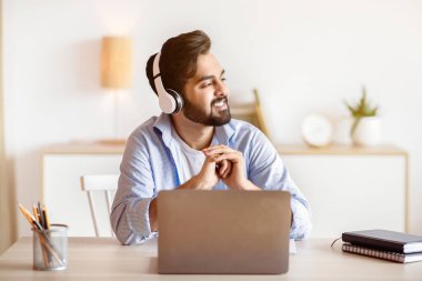 Break in Work. Ev bilgisayarında çalışırken kulaklıkla müzik dinleyen gülümseyen Arap adam, rahat bir doğulu adam bilgisayarla masada oturuyor ve bir şeyler düşünüyor.
