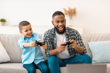 Heyecanlı Afrikalı baba ve oğlu video oyunu oynuyorlar ve evdeki oturma odasında birlikte oturup eğleniyorlar. Aile eğlencesi ve eğlencesi. Babamla Hafta Sonu