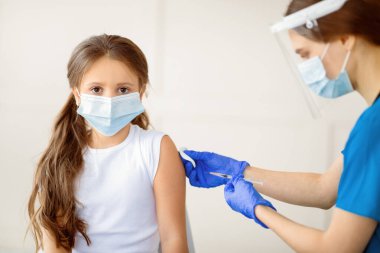 Covid-19 bağışıklık kampanyası. Yüz maskeli küçük bir hasta klinikteki hemşireden koronavirüs aşısı vuruluyor. Çocuk bulaşıcı hastalıklara karşı aşılanıyor, virüsten korunuyor.