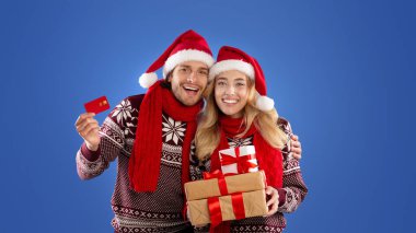 Noel alışverişi. Genç adam ve kız arkadaşı mavi arka planda bir yığın hediye ve kredi kartı ile