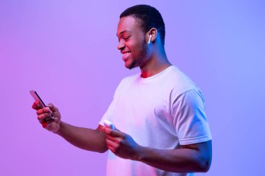 Eğlence için teknik malzemeler. Müzik dinlemek için akıllı telefon ve kulaklık kullanan, elinde kulaklık tutan neşeli Afrikalı adam Mor Arkaplan 'ın üzerinde Neon Light' ın önünde duruyor.