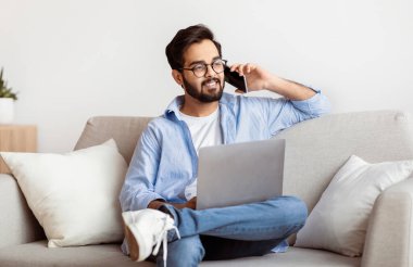 Uzak İş Fırsatları. Doğulu serbest çalışan bir adam cep telefonuyla konuşuyor ve evinde dizüstü bilgisayar kullanıyor, oturma odasında koltukta otururken bilgisayarla çalışan gülümseyen bir Arap milenyumu elemanı.