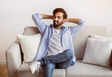 Hafta sonu evde. Rahat bir Arap adam, elleri başının arkasında koltuğa yaslanıyor, neşeli bir Orta Doğulu adam rahat bir oturma odasında dinleniyor, izin gününün keyfini çıkarıyor, boşluğu kopyalıyor.