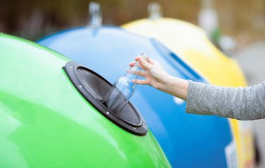 Atık tasnif ve geri dönüşüm konsepti. Tanımlanamayan bir kadın yeşil geri dönüşüm kutusuna boş cam şişe atıyor. Dışarıdaki çöp konteynırına, çevreye önem veriyor, seçici odaklı kırpılmış görüntü.