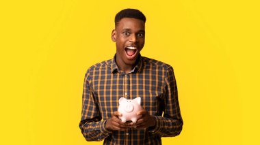 Akıllı Tasarruf. Domuz kumbaralı komik zenci adam portresi sarı stüdyo geçmişine poz veriyor duygusal Afrikalı Amerikalı adam ekonomiden keyif alıyor bütçesini ve yatırımlarını planlıyor.