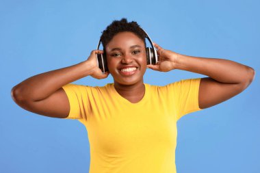 Müzik aşığı. Podcast dinleyen, kablosuz kulaklık takan, mavi arka plandaki stüdyoda poz veren, kameraya gülümseyen mutlu Afrikalı kadın. Harika Parça Listesi, Elektronik Konsepti