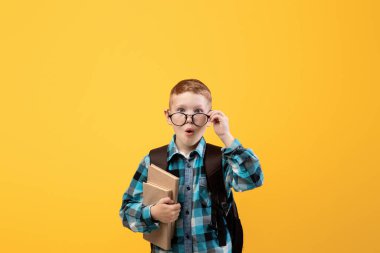 Surprised schooler with eyeglasses holding book on yellow studio background. Redhead schoolboy with backpack holding books, having amazed face expression, copy space. Books reading, education concept