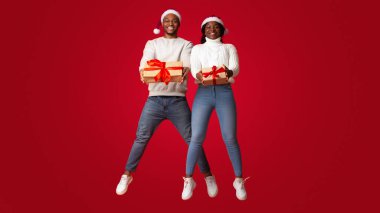 Noel Baba Şapkalı neşeli siyah çift ellerinde Noel hediyesi kutularıyla zıplıyorlar, Xmas hediyelerini tutuyorlar ve turuncu arka planda birlikte eğleniyorlar, kış tatillerini kutluyorlar,