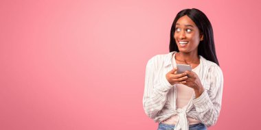 Milenyum siyahi kadın cep telefonuyla boş alana bakıyor, pembe stüdyo geçmişi var. Mutlu Afrikalı Amerikalı kadın mobil cihaz kullanıyor, internetten iletişim kuruyor, sosyal medyaya mesaj yolluyor.