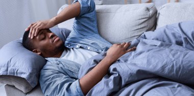 Hasta siyah adam ateşli alnına dokunuyor ve evde koltukta yatıyor. Coronavirus hastalığı, mevsimsel grip ve soğuk semptom konsepti. Covid-19 Virüs Hastalığı. Seçici Odaklanma