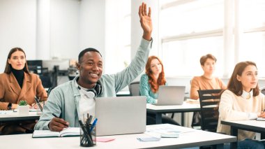 Eğitim Konsepti. Gülümseyen Afrikalı Amerikalı Erkek Öğrenci, üniversitede ders dinlerken dizüstü bilgisayarını kullanarak elini kaldırdı ve öğretim görevlisine soru sordu. Modern Parlak Öğrencilerin Çoklu Etnik Grubu