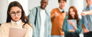 Zorbalık kavramı. Gözlüklü üzgün kadınla alay eden, dalga geçen, onu işaret eden ve gülen, bulanık arka planda duran kaba bir grup insan. Eşek şakası, taciz, toplum içinde utanç