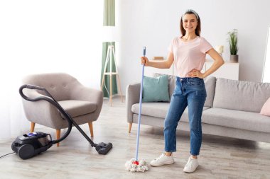 Düzenli bir dairenin ortasında paspas pozu verip gülümseyen neşeli genç hizmetçi, fotokopi odası. Pozitif temizlik hizmeti uzmanı. Sağlık hizmeti sunuyor, ev temizliği yapıyor.