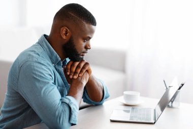 İçişleri Bakanlığı 'nda laptopla çalışan dalgın bir Afrikalı Amerikalı. Siyah serbest çalışan adam masada oturuyor ve düşünceli yüz ifadesiyle bilgisayar ekranına bakıyor, kopyalama alanı ile yan görünüm