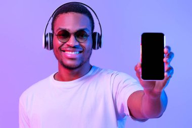 Kablosuz kulaklıklı neşeli siyah adam cep telefonuyla kamerada boş ekranı gösteriyor, Mobil Uygulama ya da Web sitesi Tasarımı için Akıllı Telefon ile Uygulama Gösteriyor, Mor Işığın Altında Duruyor