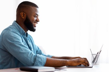 Siyah genç adam evde dizüstü bilgisayar kullanıyor, uzaktan çalışmanın tadını çıkarıyor. Gülümseyen Afrikalı Amerikalı Serbest Çalışan Adam çevrimiçi iş için bilgisayar kullanıyor, klavyede daktilo yazıyor ve ekrana bakıyor, boş alanla yan görünüm