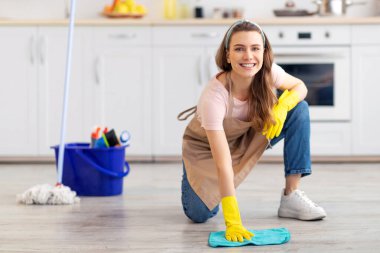 Ev idaresi konsepti. Mutfakta yerleri silen, lastik eldiven takan, temizlik malzemesi kullanan mutlu genç bir kadın. Güzel ev hanımı kameraya bakıyor ve gülümsüyor.