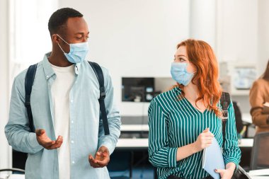 Gençlik, Yaşam Tarzı ve İnsanlar Konsepti. Bir grup uluslararası öğrenci koruyucu tıbbi maskeler takıyor ve konuşuyor, siyahi adam kızıl saçlı kadına çıkma teklif ediyor, konferans salonunda duruyor.