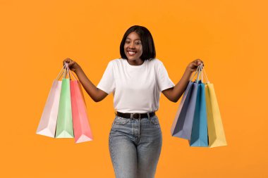 Duygusal Afrikalı Amerikalı genç kadın renkli alışveriş torbalarıyla el kaldırıyor ve gülümsüyor, satış sezonunu turuncu stüdyo arkaplanından başlayarak kutluyor.