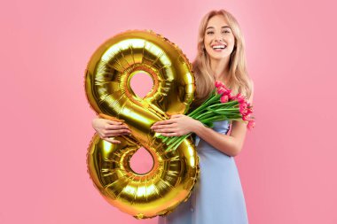 İlkbahar çiçekleri ve altın balonuyla Pembe Stüdyo Arkaplan 'da Kadın Günü' nü kutlayan çekici bir kadın. Bir sürü lale ve tatil süsü tutan güzel sarışın.