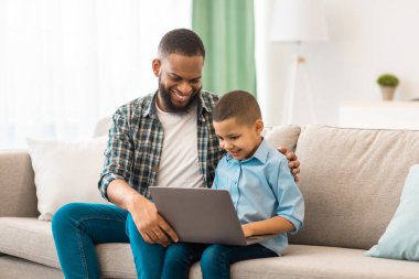 Afrika kökenli Amerikalı baba ve oğul dizüstü bilgisayar kullanarak çocuğuna bilgisayar kullanmayı ve evdeki koltukta oturarak internet izlemeyi öğretiyor. Aile Yaşamı, Teknoloji Konsepti