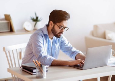 Evde masa başında oturan ve bilgisayarını daktilo eden serbest çalışan bir Arap. Yakışıklı Orta Doğulu Gözlüklü Erkek Girişimci Çevrimiçi Çalışıyor, Bilgisayar ekranına odaklanmış, Uzaktan İşlerin Tadını Çıkartıyor, Boş Alan