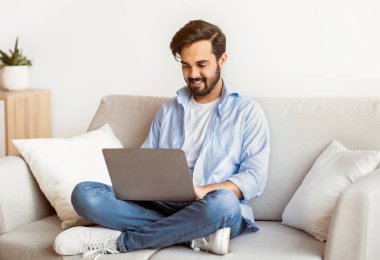 Uzak çalışma. Mutlu Arap Serbest Çalışan Adam evde dizüstü bilgisayar kullanıyor, klavyede daktilo kullanıyor, genç doğu çocuğu oturma odasında oturuyor, uzaktan internet projesi üzerinde çalışıyor, Boşluğu Kopyala