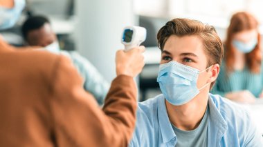 Önlem ve Önlem. Dijital tıbbi temas olmayan kızılötesi termometre tabancası kullanan kadın cerrahi maske takan erkek öğrencinin vücut ısısını ölçüyor ve tarıyor.