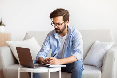 Çevrimiçi Eğitim. Doğulu genç adam dizüstü bilgisayarla çalışıyor ve not alıyor, Milenyum Arap gözlüklü adam çevrimiçi gözlük takıyor, bilgisayarla koltukta oturuyor, uzaktan öğrenmenin tadını çıkarıyor.