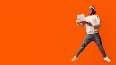 Şaşırdım, Noel Baba şapkalı siyah adam dizüstü bilgisayarla atlıyor ve heyecanla fotokopi alanına bakıyor, neşeli Afrikalı Amerikalı adam online alışverişi seviyor ve Noellerin sezonluk satışları, tam boy...