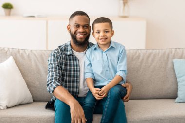 Mutlu siyah baba ve oğul portresi, evde kameraya gülümseyen babaların dizlerinin üzerinde oturan küçük çocuk. Neşeli genç baba ve çocuğu içeride poz veriyor. Aile, mutluluk ve çocukluk kavramı