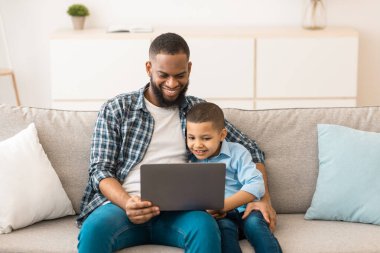 Mutlu Siyah Baba ve Oğlu evdeki koltukta oturmuş dijital tablet kullanarak interneti tarıyor. Küçük çocuk ve babası hafta sonları internetten film izliyorlar. Aile Eğlencesi ve Malzemeler