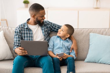 Afrikalı baba ve küçük oğlu dijital tablet bilgisayar kullanıyor evde internet tarıyor, evdeki koltukta oturuyor. Gadget ve Aile Hafta Sonu. Baba ve Çocuk İnternette Film İzliyor