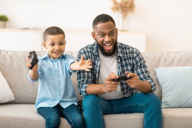 Video oyunları. Afrikalı Amerikalı baba ve oğlu evde video oyunu oynarken eğleniyorlar. Genç Baba ve Küçük Çocuk Hafta Sonu Bilgisayar Oyunları Konsepti