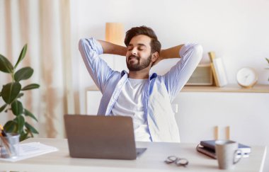 Başarılı çalışma günü. Yakışıklı Arap işadamı, elleri arkada, koltuğuna yaslanmış Orta Doğulu erkek girişimcinin çalışma masasında dinlenmesine seviniyor.